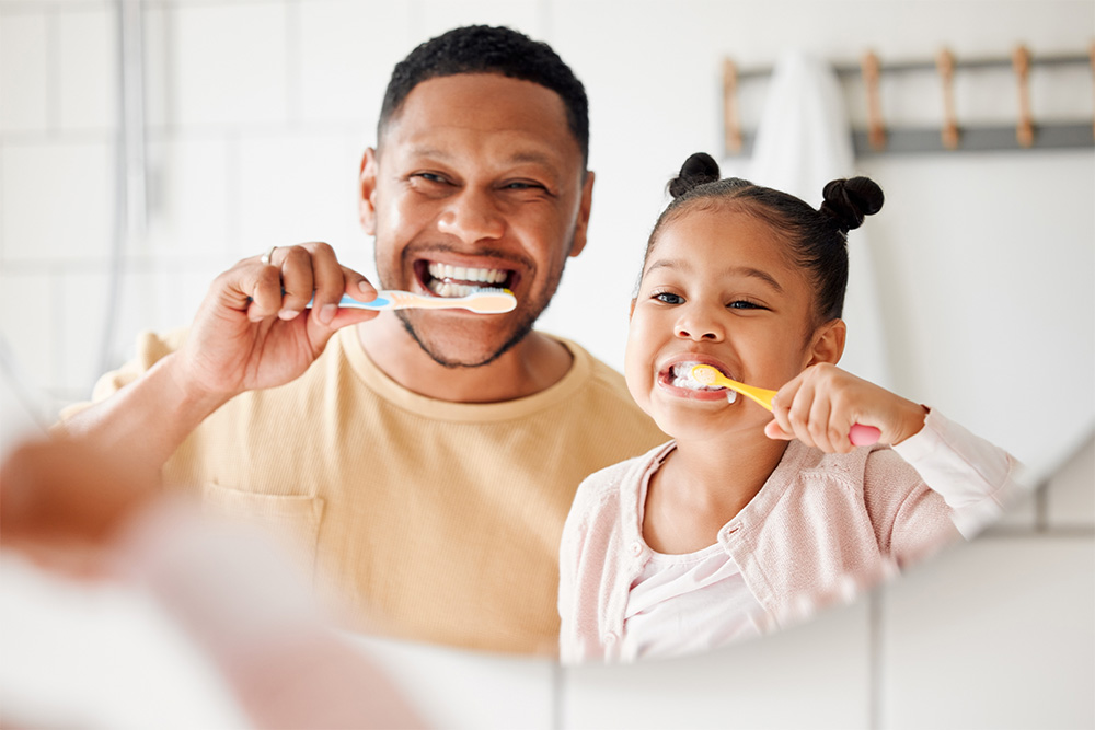 A man and a little girl happily brushing their teeth together in a bright bathroom.