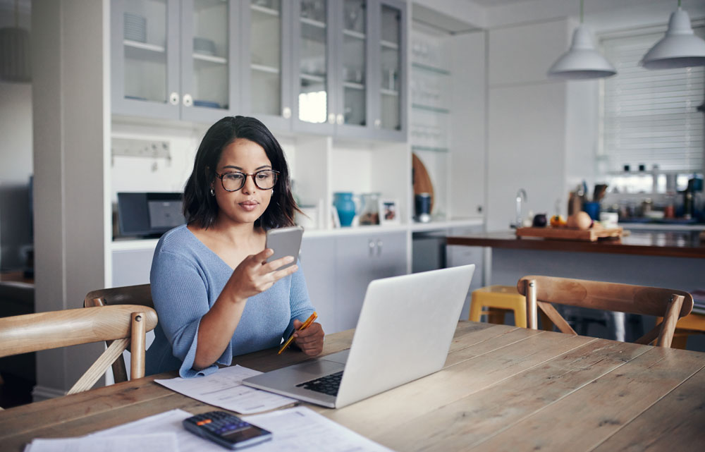 woman sitting at kitchen table looking at phone and computer