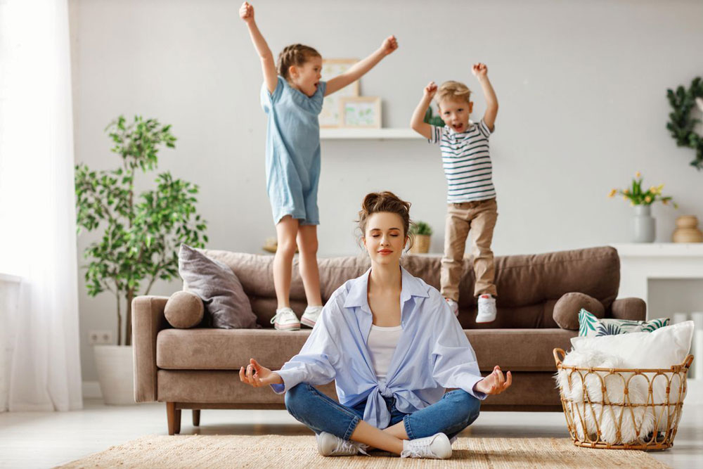 woman cross legged on floor while kids jump on sofa